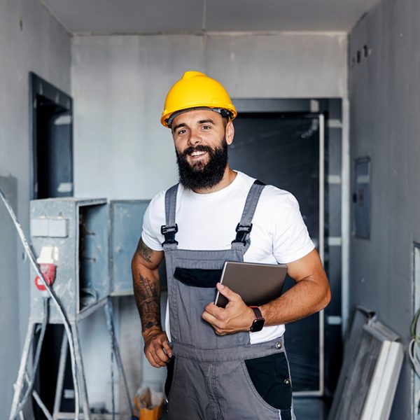 A happy construction worker with a helmet is standing in an unfinished building and holding a tablet under the armpit. He is checking on works on site.